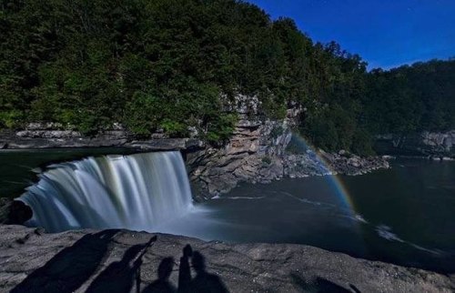 A serene waterfall cascades into a river, surrounded by lush trees, with a faint rainbow visible in the mist.
