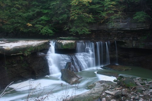 A serene waterfall cascades over rocky ledges, surrounded by lush green trees and a tranquil river below.