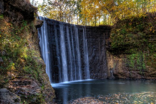 A serene waterfall cascading over rocks into a calm pool, surrounded by lush greenery and autumn foliage.