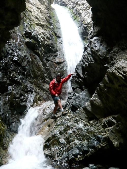 A person in a red jacket climbs a rocky wall beside a waterfall in a narrow canyon. Water cascades down the rocks.