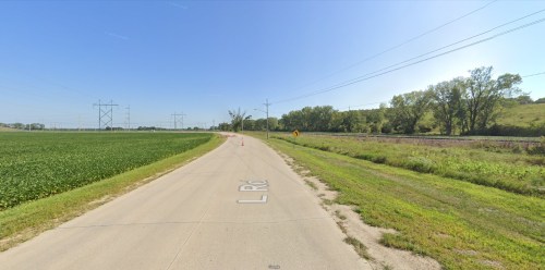 A rural road curves through green fields, with power lines and a railway visible in the background under a clear blue sky.