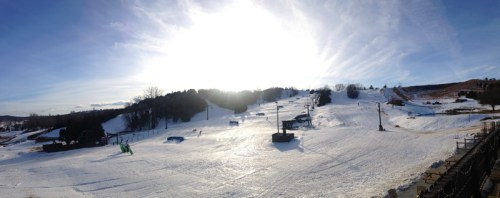 A panoramic view of a snowy ski slope under a bright sun, with ski lifts and trees in the background.