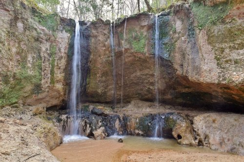 A serene waterfall cascading over rocky cliffs into a sandy area, surrounded by lush greenery and natural rock formations.