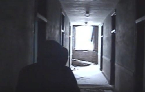 A dimly lit hallway with a figure in a hooded jacket, snow visible outside through an open door at the end.