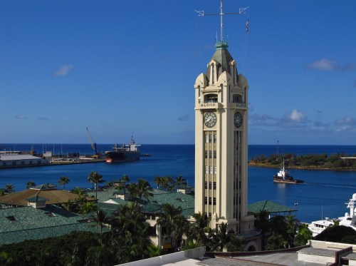 A tall clock tower stands by the harbor, surrounded by palm trees and blue waters under a clear sky.