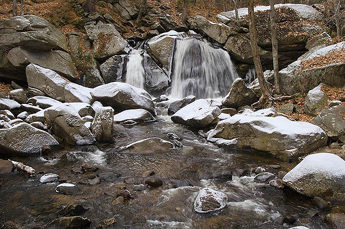A serene waterfall cascades over rocks, surrounded by snow-dusted boulders and autumn leaves in a tranquil forest setting.