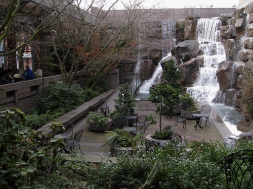A serene garden with a waterfall, surrounded by greenery and outdoor seating.