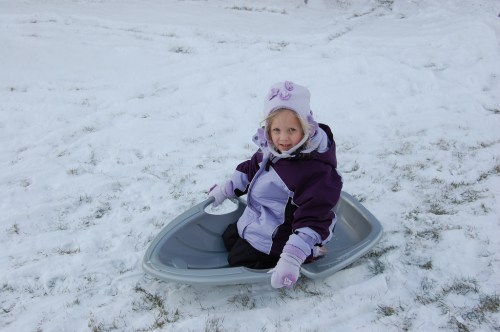 A young girl in winter clothing sits in a plastic sled on snowy ground, smiling at the camera.
