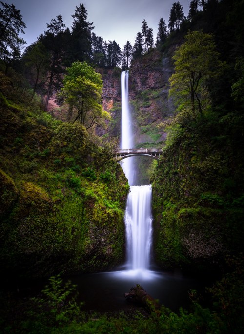 A stunning waterfall cascades between lush green cliffs, with a wooden bridge arching above the lower falls.