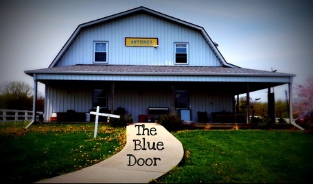 A rustic building with a sign reading "Antiques" and a path leading to a blue door. Green grass surrounds the entrance.