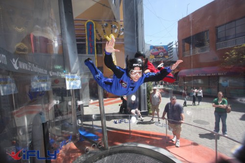 A person in a blue jumpsuit is indoor skydiving in a wind tunnel, with an instructor nearby.