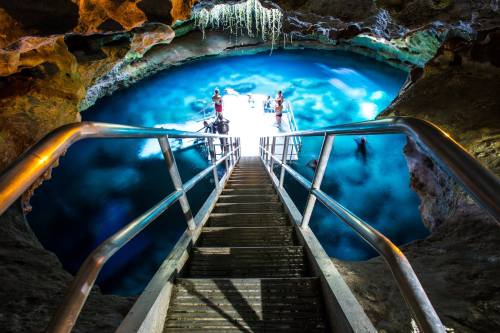 A wooden staircase leads down to a bright blue underground pool surrounded by rocky cave walls.