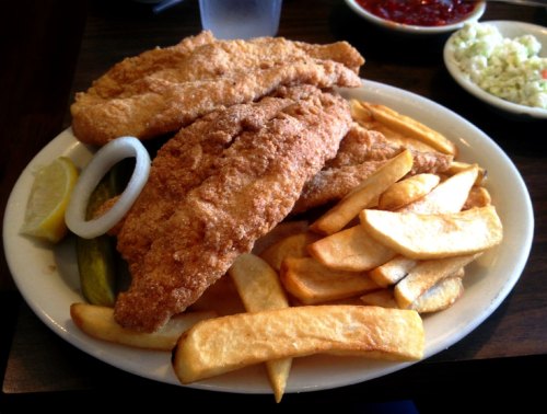 A plate of crispy fried fish fillets served with golden fries, lemon wedges, pickles, and a side of coleslaw.