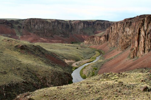 A winding river flows through a rugged canyon with steep, colorful rock formations and sparse vegetation.
