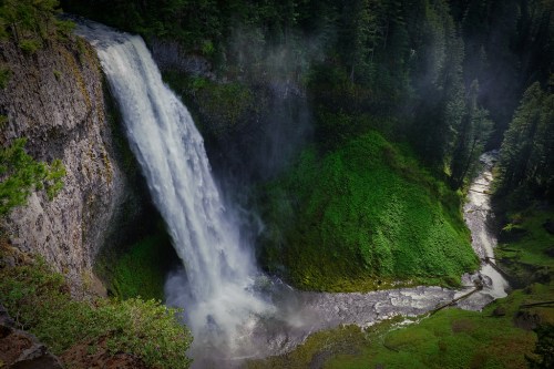 A majestic waterfall cascades down rocky cliffs, surrounded by lush green vegetation and mist in a serene forest setting.