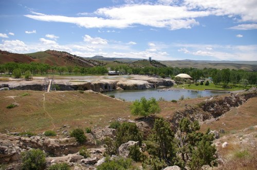 A scenic landscape featuring a calm lake, green trees, and rolling hills under a blue sky with scattered clouds.