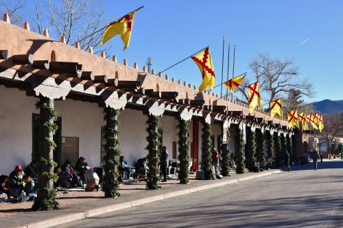 A street scene with adobe buildings, festive flags, and people sitting along the sidewalk under a clear blue sky.