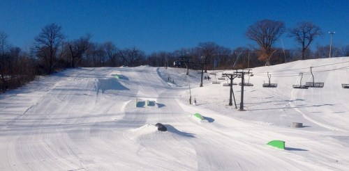 A snowy ski slope with green ramps and a ski lift under a clear blue sky.