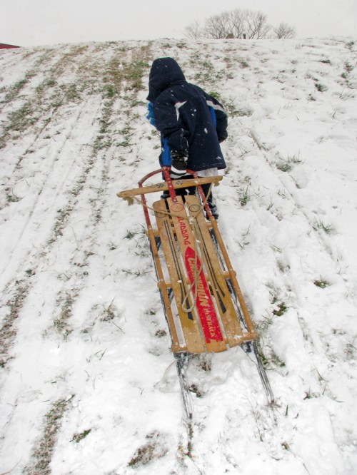 A child in a blue jacket pulls a wooden sled up a snowy hill on a winter day. Snowflakes are falling around them.