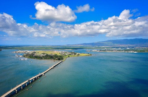 Aerial view of a coastal area with a bridge, green land, and blue waters under a partly cloudy sky.