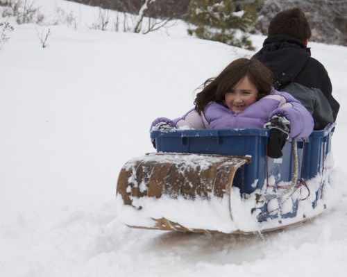 A child smiles while riding in a sled down a snowy hill, with another person steering from behind.