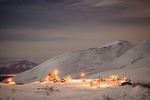 A snowy landscape at night, featuring illuminated buildings and mountains in the background under a starry sky.