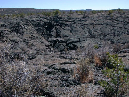 A rugged landscape of cracked black lava rock, with sparse vegetation and distant hills under a clear blue sky.