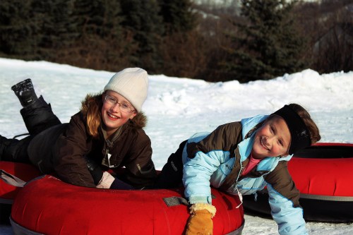Two children smiling while lying on red snow tubes in a snowy landscape, wearing winter clothing and hats.