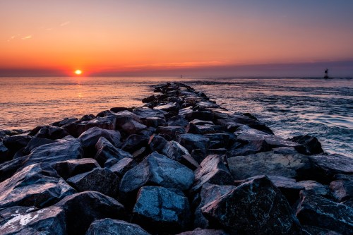 A rocky jetty extends into the ocean at sunset, with vibrant colors reflecting on the water.
