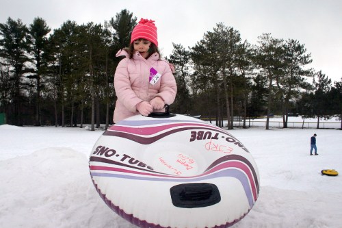 A girl in a pink coat and hat stands on a snow tube, ready to slide down a snowy hill, with trees in the background.
