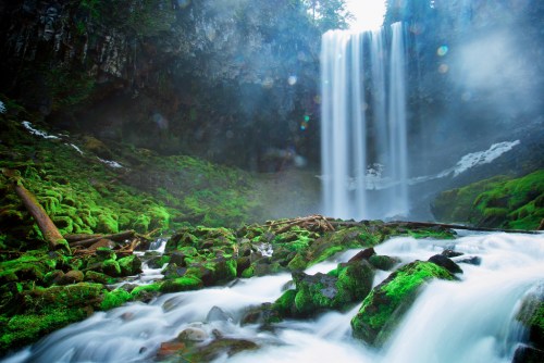 A serene waterfall cascades over moss-covered rocks, surrounded by lush greenery and mist in a tranquil natural setting.
