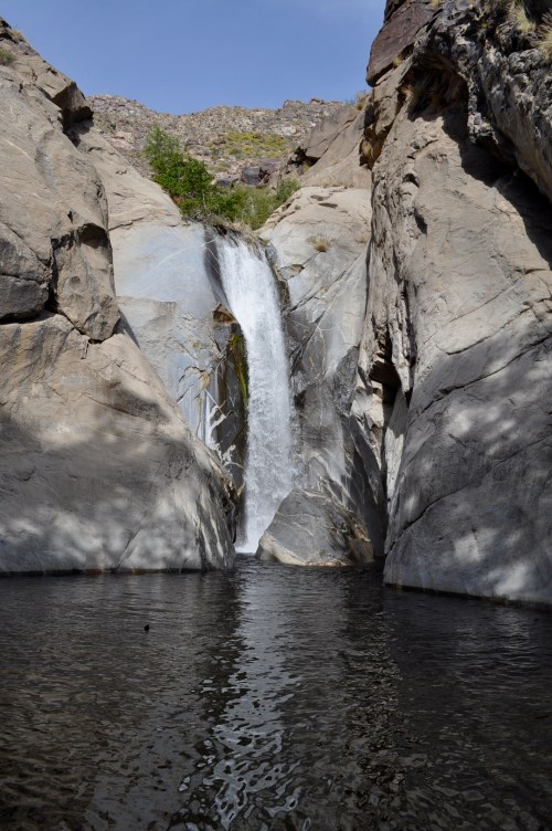A waterfall cascades down rocky cliffs into a serene pool, surrounded by rugged terrain and clear blue skies.