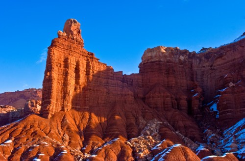 A towering red rock formation against a clear blue sky, showcasing rugged textures and shadows in a desert landscape.