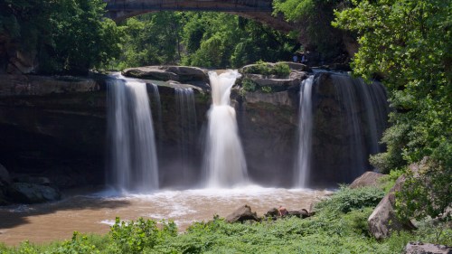 A scenic waterfall cascades over rocks, surrounded by lush greenery and a stone bridge in the background.