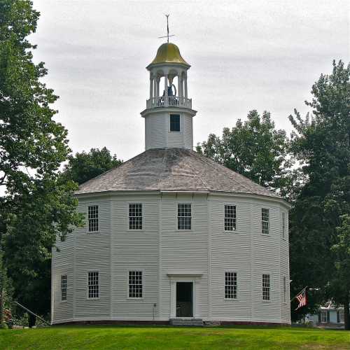 A round white church with a golden dome and a steeple, surrounded by trees and grass.