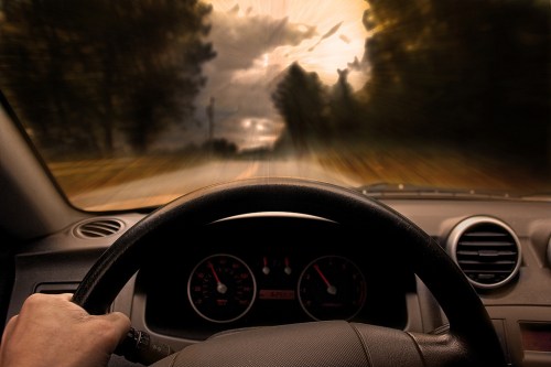 A driver's hand on the steering wheel, with a blurred view of a road and dramatic clouds in the background.