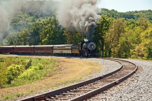 A steam train chugs along a curved track, billowing smoke against a backdrop of green trees and hills.