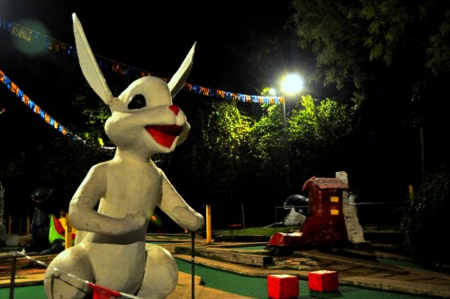 A large, white rabbit statue with red accents sits in a dimly lit mini-golf course, surrounded by colorful decorations.