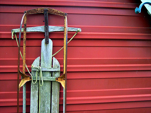 An old wooden sled leaning against a red barn wall, with rusty metal parts and worn-out wood.