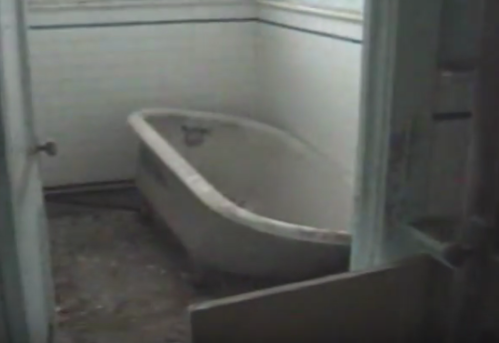 An old, empty bathtub in a dusty, abandoned bathroom with white tiled walls.