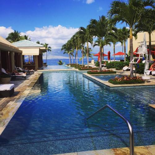 A serene resort pool surrounded by palm trees, lounge chairs, and a view of the ocean under a partly cloudy sky.