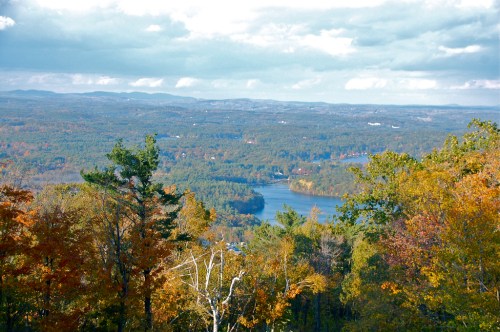 A scenic view of a river surrounded by autumn foliage and rolling hills under a cloudy sky.