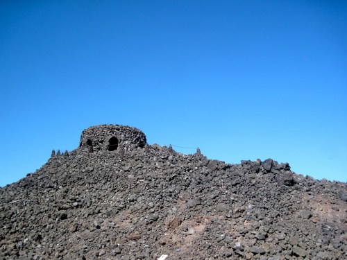 A rocky hill topped with a stone structure against a clear blue sky.