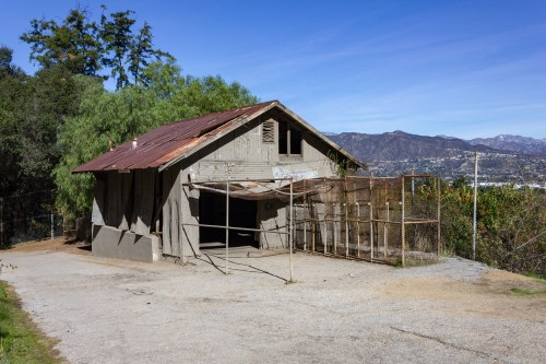 A weathered house with a rusty roof, surrounded by trees and mountains, set against a clear blue sky.