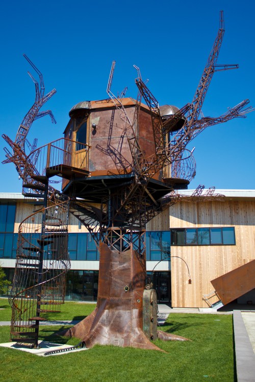 A large, rusted metal treehouse with a spiral staircase, set against a clear blue sky and modern building backdrop.