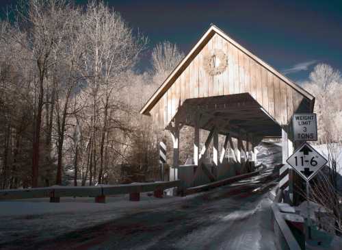 A snow-covered covered bridge with a wooden structure and a wreath, surrounded by bare trees and a weight limit sign.