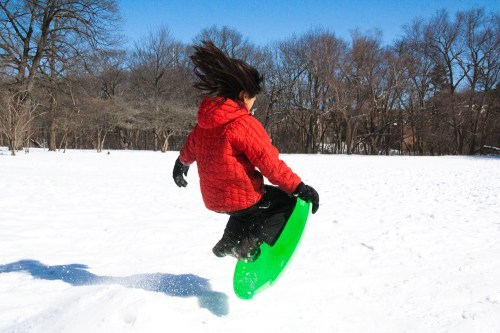 A child in a red jacket jumps on a green sled, soaring through the snow on a sunny winter day.