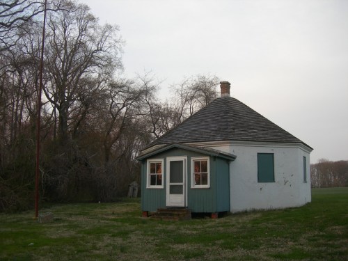 A small, round house with a sloped roof, set in a grassy area surrounded by trees.