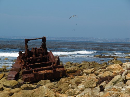 Rusty machinery sits on rocky shore with waves and seagulls in the background under a clear blue sky.