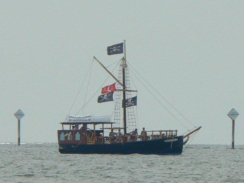 A pirate ship with black flags sails on calm waters, surrounded by navigational markers in the distance.